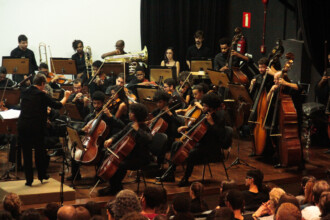 A imagem mostra uma orquestra em um palco escuro, com um regente de costas para a câmera em primeiro plano. O regente, que usa roupas pretas, está de pé em um púlpito, gesticulando para a orquestra. A orquestra, que é composta por cerca de 20 a 30 músicos, está tocando instrumentos de corda, como violoncelos e contrabaixos. No fundo do palco, há uma cortina preta e alguns músicos tocando instrumentos de sopro, como um trombone. A audiência, vista do ponto de vista do palco, é visível na parte inferior da imagem.