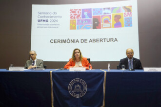 A imagem mostra três pessoas sentadas em uma mesa em um palco, com um telão ao fundo. No centro, uma mulher de cabelos loiros e casaco laranja está sentada à frente. À sua direita, um homem de terno cinza e óculos está sentado; à sua esquerda, um outro homem de terno escuro e careca. Os três estão com as mãos apoiadas sobre a mesa, que está coberta por uma toalha azul. Na parte da frente da mesa, um pano azul com o brasão da Universidade Federal de Minas Gerais (UFMG) está pendurado. No telão, ao fundo, há um slide com o título "Semana do Conhecimento UFMG 2024", seguido pela frase "Diversidade: conhecer, preservar e restaurar". Ao lado, uma ilustração colorida exibe diversas formas e perfis de rostos. Abaixo, a frase em destaque "CERIMÔNIA DE ABERTURA" está escrita em letras grandes e cinzas. No canto inferior direito da imagem, uma pequena garrafa de álcool em gel pode ser vista sobre a mesa.