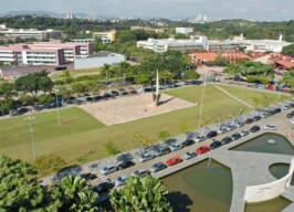 A imagem é uma vista aérea de um campus universitário com áreas verdes, edifícios e estacionamentos. No centro da imagem, há um grande gramado com um monumento de metal no centro, cercado por um caminho pavimentado. À esquerda e à direita do gramado, há longas fileiras de carros estacionados em vagas ao ar livre. Ao fundo, uma série de edifícios de diferentes cores e tamanhos se estendem, incluindo um prédio cor-de-rosa no lado esquerdo. Árvores e vegetação preenchem as áreas entre os edifícios. Na parte inferior direita, um espelho d'água de formato irregular é visível. Ao longe, uma paisagem de cidade é vista no horizonte.