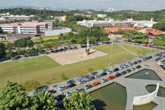 A imagem é uma vista aérea de um campus universitário com áreas verdes, edifícios e estacionamentos. No centro da imagem, há um grande gramado com um monumento de metal no centro, cercado por um caminho pavimentado. À esquerda e à direita do gramado, há longas fileiras de carros estacionados em vagas ao ar livre. Ao fundo, uma série de edifícios de diferentes cores e tamanhos se estendem, incluindo um prédio cor-de-rosa no lado esquerdo. Árvores e vegetação preenchem as áreas entre os edifícios. Na parte inferior direita, um espelho d'água de formato irregular é visível. Ao longe, uma paisagem de cidade é vista no horizonte.