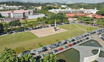 A imagem é uma vista aérea de um campus universitário com áreas verdes, edifícios e estacionamentos. No centro da imagem, há um grande gramado com um monumento de metal no centro, cercado por um caminho pavimentado. À esquerda e à direita do gramado, há longas fileiras de carros estacionados em vagas ao ar livre. Ao fundo, uma série de edifícios de diferentes cores e tamanhos se estendem, incluindo um prédio cor-de-rosa no lado esquerdo. Árvores e vegetação preenchem as áreas entre os edifícios. Na parte inferior direita, um espelho d'água de formato irregular é visível. Ao longe, uma paisagem de cidade é vista no horizonte.