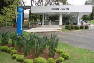 A imagem mostra uma vista externa de um edifício de um andar, com fachada branca e grandes janelas de vidro. O letreiro na parte superior do prédio exibe as letras "CNEN - CDTN" em preto. Uma escadaria de concreto com corrimões leva à entrada do edifício. Em primeiro plano, há um canteiro de jardim com folhagens verdes escuras e arbustos redondos, cercado por gramado. À esquerda do canteiro, uma placa de sinalização azul e cilíndrica com a inscrição "Bem-vindo / Welcome" em branco está ao lado de uma estrutura de entrada.