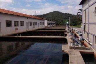 A imagem mostra uma estação de tratamento de água em um ambiente externo. No centro, tanques retangulares, de concreto e cheios de água, se estendem em fila. Equipamentos e tubulações de metal estão instalados ao longo da lateral direita dos tanques. Ao fundo, há prédios brancos com telhados de telhas vermelhas e janelas em suas fachadas. O ambiente é iluminado pela luz do sol e o céu está claro, com nuvens brancas. Ao longe, uma colina coberta por mata verde é visível no horizonte.