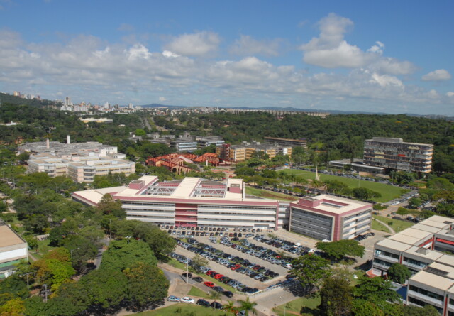 L'image montre une vue grand angle en hauteur d'un vaste campus universitaire par une journée ensoleillée. Au premier plan, un grand bâtiment moderne à la façade rouge et blanche domine la vue, avec un immense parking bondé juste devant. Le campus est très verdoyant, avec de nombreux arbres et des parcelles de forêt entrecoupées entre les différents bâtiments universitaires. Au-delà du bâtiment central, on distingue d'autres installations du campus, dont un terrain de sport et plusieurs autres bâtiments à plusieurs étages aux styles architecturaux variés. La silhouette de la ville est visible au loin sur la gauche du cadre, suggérant que l'université est située dans ou en périphérie d'une grande agglomération. Le ciel est d'un bleu éclatant parsemé de nuages ​​blancs. L'impression générale est celle d'une grande institution bien établie, nichée dans un paysage vallonné et luxuriant.