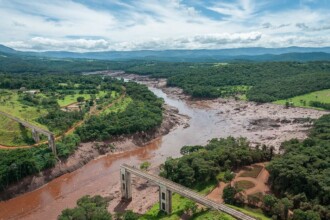 A imagem, vista de uma perspectiva elevada, mostra uma paisagem de uma área rural com um rio de água e lama de cor marrom que a atravessa. A terra ao redor do rio é de cor escura, contrastando com a vegetação verde que cobre a maior parte da paisagem. Há duas pontes sobre o rio, que parecem ser de uma ferrovia, uma em primeiro plano e outra mais ao fundo à esquerda. As colinas verdes ao longe se estendem até o horizonte, sob um céu com nuvens brancas e esparsas.