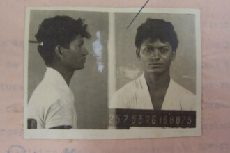 A imagem é de uma fotografia em preto e branco, que contém duas fotos de um homem jovem, lado a lado. A fotografia está sobre uma folha de papel rosa, que contém textos e rabiscos. O homem, de cabelo escuro e com volume, usa uma camisa de cor clara. A foto da esquerda mostra o perfil do rosto do homem, olhando para a direita. A foto da direita mostra o rosto dele de frente. Na parte de baixo da foto, há um cartão de identificação com um número de registro, que está desfocado.