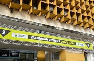 A imagem mostra a fachada de um prédio de vários andares, com uma estrutura de janelas quadradas e recuadas, formando um padrão de favo de mel. O prédio é de cor amarela mostarda e está em um dia claro, com o céu azul e algumas nuvens. Na frente do prédio, há um grande banner triangular com a inscrição "VACINAÇÃO COVID-19 DRIVE-THRU", com o logotipo da prefeitura. O banner é sustentado por uma estrutura metálica e indica "2" à esquerda e "1" à direita. Em primeiro plano, mesas brancas de plástico e um cavalete de plástico estão presentes, e uma pessoa está em pé ao lado de um balcão, no canto direito.
