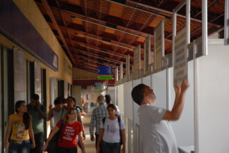 A imagem mostra a vista da entrada principal de um prédio com fachada em tons de bege e janelas com grades e esquadrias marrons. Uma rampa e uma escada com corrimãos levam até a entrada do edifício. Em primeiro plano, árvores de grande porte, com troncos largos e copas frondosas, se destacam. Ao lado da rampa, há duas motos estacionadas e uma bicicleta.