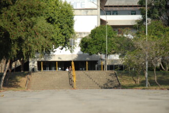 A imagem mostra um auditório de uma universidade, com cadeiras em semicírculo e um homem em uma cadeira de rodas no lado esquerdo do quadro. O homem está de perfil, usando uma camisa cinza e calças escuras, e gesticula com as mãos enquanto fala. A maioria das pessoas está de costas para a câmera, prestando atenção ao homem, enquanto uma mulher em primeiro plano, com cabelos curtos e óculos de sol na cabeça, está de perfil com a mão no rosto. O ambiente tem paredes brancas, e as cadeiras são de cor escura.