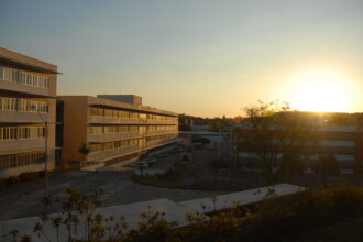 A imagem mostra a vista externa de um conjunto de edifícios em um campus, com o pôr do sol no horizonte. No centro da imagem, há um prédio de três andares de cor bege, com longas fileiras de janelas. As janelas, com venezianas, refletem a luz do sol. Um estacionamento com carros é visível na frente do edifício. À esquerda, um outro prédio de três andares, com paredes de cor clara e varandas com grades brancas, está parcialmente visível. À direita, um edifício vermelho com janelas grandes é iluminado pelo sol, que está se pondo no horizonte. A luz amarelada e dourada do sol ilumina a paisagem, criando um brilho intenso no céu claro.