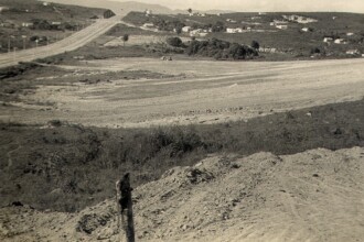A imagem em preto e branco mostra uma paisagem de terreno aberto e em declive. A cena é dominada por solo exposto e áreas de vegetação rasteira. Em primeiro plano, há um monte de terra com um toco de madeira. No plano intermediário, há um trecho de estrada de terra visível e, mais ao fundo, a topografia se eleva com casas esparsas e vegetação. Do lado esquerdo, no topo, uma estrada pavimentada com postes de luz segue a curva da colina, desaparecendo no horizonte.