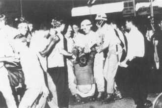 A imagem em preto e branco mostra um aglomerado de pessoas em um ambiente externo à noite. A cena é caótica, com várias pessoas em pé e uma pessoa no centro do grupo sendo carregada por outras. A iluminação é esparsa, e o foco da imagem está no grupo central de pessoas. Algumas pessoas usam capacetes brancos, enquanto outras estão vestidas com roupas claras e escuras.