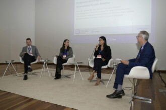 A imagem mostra um painel de discussão com quatro pessoas em um palco. As pessoas estão sentadas em cadeiras brancas, de frente para a câmera. Um homem de terno cinza, uma mulher de blazer escuro, uma mulher de blusa e casaco escuros, e um homem de terno azul estão na ordem da esquerda para a direita. O homem da esquerda está segurando um microfone. Ao fundo, um telão grande exibe o título de um evento e os nomes e títulos dos participantes, incluindo o nome "Alexandre Morais", "Vice-reitor da Universidade Federal de Minas Gerais". Pequenas mesas redondas, com copos e garrafas d'água, estão dispostas entre as cadeiras. O palco tem um chão de madeira e um tapete branco.