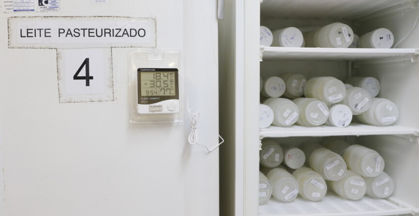 imagem mostra um refrigerador branco em um ambiente interno. A porta do refrigerador à esquerda tem uma placa com o texto "LEITE PASTEURIZADO" e o número "4". Um termômetro digital está fixado na porta, exibindo as temperaturas "18.4°C" e "-30.5°C". A porta do refrigerador está aberta, revelando seu interior com várias prateleiras. As prateleiras estão cheias de potes ou frascos de plástico, brancos e transparentes, dispostos lado a lado. A iluminação é difusa, e o ambiente parece ser um laboratório ou um espaço de armazenamento.