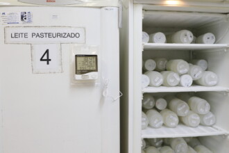 imagem mostra um refrigerador branco em um ambiente interno. A porta do refrigerador à esquerda tem uma placa com o texto "LEITE PASTEURIZADO" e o número "4". Um termômetro digital está fixado na porta, exibindo as temperaturas "18.4°C" e "-30.5°C". A porta do refrigerador está aberta, revelando seu interior com várias prateleiras. As prateleiras estão cheias de potes ou frascos de plástico, brancos e transparentes, dispostos lado a lado. A iluminação é difusa, e o ambiente parece ser um laboratório ou um espaço de armazenamento.