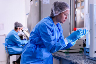 A imagem mostra uma mulher em um laboratório, vista de lado e de frente. Ela está vestindo um jaleco azul, luvas azuis e uma touca de cabelo branca. Ela segura um frasco transparente, que tem uma tampa vermelha, e está olhando para ele com atenção. Ela está perto de um equipamento branco de metal. No braço esquerdo, uma parte da mão dela está levantando a tampa do frasco. Ao fundo, outra pessoa, também com jaleco azul, luvas e touca, está sentada em uma cadeira, olhando para um microscópio. O ambiente do laboratório é claro, com bancadas de cor escura, equipamentos científicos, e paredes brancas.