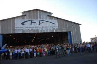 A imagem mostra uma vista externa de um grande hangar de madeira, com o portão principal aberto. Na fachada do hangar, um grande banner com o logotipo "CEA" e o texto "Centro de Estudos Aeronáuticos - Escola de Engenharia" e "UNIVERSIDADE FEDERAL DE MINAS GERAIS" está fixado acima da entrada. O céu, visível no topo da imagem, está claro, e um avião branco pode ser visto voando ao longe, à direita. Uma grande multidão de pessoas está em frente ao hangar, assistindo a um evento. No meio do grupo, um grupo de jovens uniformizados está em destaque. Em primeiro plano, uma parte de um avião azul, com as asas brancas, é visível à esquerda. Ao fundo, uma banda com instrumentos de sopro está presente no meio da multidão. A iluminação é natural, proveniente da luz do dia, criando uma atmosfera clara e iluminada.