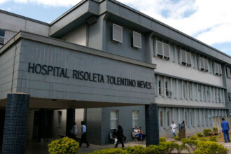 A imagem mostra uma vista externa de um edifício de cor cinza, com três andares. Na parte da frente do prédio, um letreiro grande com letras pretas exibe os dizeres "HOSPITAL RISOLETA TOLENTINO NEVES". O prédio tem uma entrada com uma marquise, sustentada por colunas cinzas cilíndricas. Em frente à entrada, algumas pessoas estão caminhando e outras estão sentadas em um banco. Uma vegetação baixa e alguns arbustos estão plantados ao redor do edifício. A iluminação é natural, proveniente da luz do dia, e o céu, visível no canto superior direito, tem algumas nuvens.