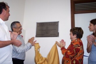 A imagem mostra quatro pessoas adultas em pé, em um ambiente interno. Elas estão batendo palmas em frente a uma placa metálica fixada em uma parede branca. No centro da cena, dois tecidos, um de cor dourada e outro azul, estão sendo removidos da placa. A placa de inauguração é retangular e está no centro da parede, com um texto gravado. À esquerda, um homem de camisa branca e outro com camisa social cinza estão batendo palmas. À direita, uma mulher com uma blusa estampada em tons de laranja e marrom e outra mulher de blusa azul também estão aplaudindo. A iluminação é proveniente do interior do ambiente.