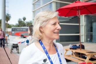 A imagem mostra o retrato de uma mulher de cabelos loiros e curtos, vista em perfil, sorrindo e olhando para a direita. Ela usa brincos de pérolas, uma camiseta branca com um cordão de pescoço azul e branco, e um cordão com uma medalha. A camiseta tem o logotipo da Adidas e, abaixo, o logotipo dos Jogos Paralímpicos. O fundo da imagem é um espaço externo com um prédio de fachada de vidro e uma grande marquise vermelha. Um ônibus vermelho e cinza e pessoas, incluindo uma pessoa em uma cadeira de rodas, são visíveis em segundo plano, no lado esquerdo da imagem. No canto inferior direito, uma mesa de madeira e um banco também são visíveis.