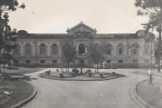 A imagem em preto e branco mostra um edifício de arquitetura clássica com vários andares e janelas, em um estilo simétrico. O prédio tem uma entrada central com uma varanda e uma escadaria, e uma ornamentação acima da entrada principal. No primeiro plano, há um canteiro central circular com arbustos, rodeado por uma rua em forma de Y que se bifurca em direção aos lados. À direita, um homem vestido com camisa clara e calça escura caminha na calçada. Em frente ao prédio, um carro antigo está estacionado. Duas árvores, uma de cada lado, emolduram a imagem.