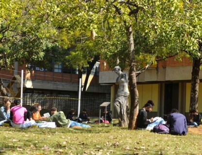 A imagem mostra um grupo de pessoas sentadas na grama, em um ambiente externo, à sombra de árvores. No primeiro plano, à esquerda, quatro pessoas estão sentadas em círculo, conversando e lendo. Outras pessoas estão sentadas individualmente ou em pequenos grupos. A pessoa no canto inferior esquerdo está de camisa rosa, sentada de frente para as outras. Entre as árvores, uma estátua escura de uma figura feminina está visível. O chão é coberto por grama verde, com algumas folhas secas. Ao fundo, um prédio de tijolos vermelhos e grandes janelas retangulares é visível. A luz do sol é intensa e cria áreas de sombra.