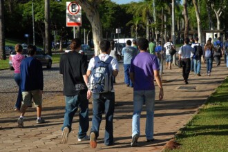 A imagem mostra um grupo de pessoas caminhando em uma calçada de blocos de cimento em um ambiente externo. No primeiro plano, três jovens, vistos de costas, estão caminhando em direção ao centro da imagem. O do meio usa uma mochila azul e branca. Ao fundo, há um estacionamento com carros, cercado por grama e árvores. Outras pessoas, em grupos e sozinhas, estão caminhando na mesma direção. Uma placa de trânsito branca, com o símbolo de "E" cortado, indicando proibido estacionar, está visível no canto superior esquerdo. A iluminação é natural, proveniente da luz do sol.