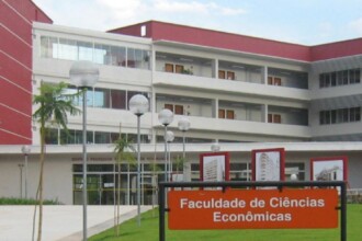 A imagem mostra uma vista externa de um edifício de dois andares com fachada de cor vermelha e branca. Há um caminho que leva a uma entrada principal com portas de vidro. Um letreiro grande e laranja, em primeiro plano, indica "Faculdade de Ciências Econômicas". Ao longo do caminho, há postes de iluminação brancos e redondos, e pequenas árvores. A paisagem é complementada por um gramado verde, árvores e um céu azul com nuvens brancas e esparsas.
