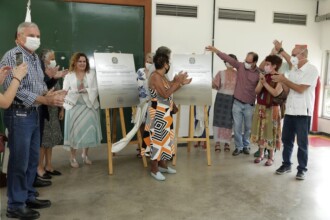 A imagem mostra um grupo de pessoas em um ambiente interno, celebrando e aplaudindo a inauguração de algo. No centro, duas placas de metal estão em cavaletes de madeira. Uma mulher com um vestido estampado em preto, branco e laranja está à frente das placas, aplaudindo. Ao seu redor, as outras pessoas aplaudem, algumas com as mãos levantadas, enquanto a maioria usa máscaras de proteção facial brancas. O cenário é uma sala com uma parede verde na parte superior e branca na parte inferior.