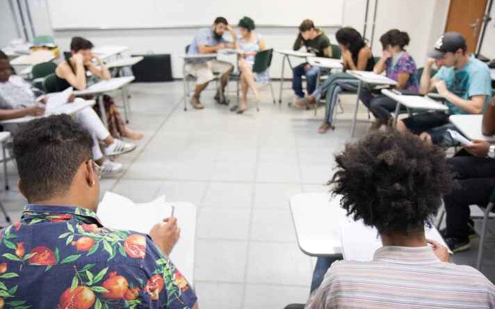 Imagem mostra sala com diversas pessoas sentadas em carteiras escolares, dispostas em forma de um círculo