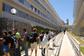 A imagem mostra uma vista externa de um complexo de edifícios de cor clara, com uma rampa de acesso no primeiro plano. A rampa, com um corrimão branco, leva a uma passarela que segue paralela ao prédio principal. Várias pessoas estão caminhando ao longo da rampa, em direção ao fundo. À esquerda da rampa, há um grupo de jovens conversando, alguns com mochilas nas costas. Um muro de pedras está presente ao lado da rampa, e um poste de luz com uma luminária redonda e branca está no topo. Ao fundo, o prédio de vários andares, com longas fileiras de janelas, se estende por toda a imagem. A luz do sol é intensa, e o céu está azul e claro.