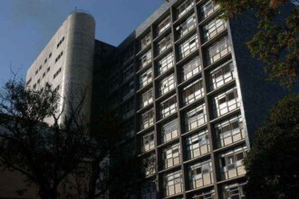 A imagem mostra uma vista angular de um edifício alto. A fachada principal do prédio é retangular e cinza, com várias janelas retangulares dispostas em um padrão de grade. A maioria das janelas é de vidro e as persianas de metal estão visíveis. No canto superior esquerdo do prédio, há uma estrutura cilíndrica, também de cor cinza e de vários andares. Algumas árvores e seus galhos, com folhas escuras, estão em primeiro plano. O céu é azul e iluminado, mas a luz do sol não é intensa.