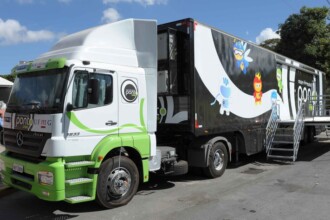 A imagem mostra a lateral de um caminhão-baú que foi adaptado para ser uma unidade móvel. O caminhão tem a cabine branca com detalhes verdes e o nome "Ponto UFMG" e "Mercedes-Benz" visíveis na lateral. A placa do veículo está visível no para-choque dianteiro. A parte traseira do caminhão é um baú grande e preto, com uma grande faixa branca na lateral e diversas ilustrações de personagens de desenho animado. Ao final do baú, uma escada metálica com corrimão foi montada, servindo de acesso para a parte interna. O caminhão está estacionado em uma rua asfaltada.
