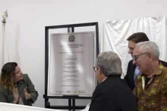 A imagem mostra quatro pessoas em um ambiente interno. No centro, uma placa de metal retangular com texto gravado está em um cavalete preto. A placa diz "REPÚBLICA FEDERATIVA DO BRASIL", "UNIVERSIDADE FEDERAL DE MINAS GERAIS", "ESCOLA DE BELAS ARTES", seguido pela data "Belo Horizonte, 28 de junho de 2024". Dois homens estão em pé à direita, um de terno azul escuro e outro com uma camisa estampada em tons de marrom e amarelo, olhando e rindo para a placa. À esquerda, uma mulher de blusa verde está batendo palmas. Um homem de terno cinza, de costas, está em frente à placa. A parede é branca, e um mastro com uma bandeira branca pode ser visto no canto esquerdo.