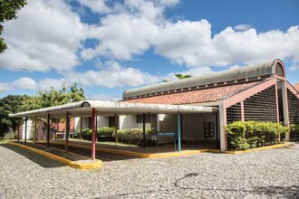 A imagem mostra um edifício de fachada branca com telhado inclinado de telhas marrons. Uma estrutura de concreto e metal, que lembra um toldo, é visível na parte inferior do prédio. A área em frente ao edifício é pavimentada com paralelepípedos, e o toldo é sustentado por pilares de cores variadas, como amarelo, azul, branco e vermelho. Um grande canteiro com arbustos verdes e uma calçada com grama completam a cena. O fundo da imagem é preenchido por um céu com nuvens brancas e esparsas.