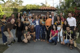 A imagem mostra um grupo de 25 pessoas, incluindo homens e mulheres, posando para uma foto em um pátio externo. O grupo está organizado em duas fileiras, uma em pé e outra agachada na frente. Algumas pessoas usam adornos culturais, como cocares, pintura facial e colares. O cenário é um ambiente externo com vegetação exuberante e uma parede de madeira e telhas ao fundo. Todos no grupo olham em direção à câmera, com expressões neutras.