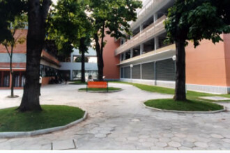 A imagem mostra um pátio externo entre edifícios, pavimentado com blocos de concreto hexagonais. O ambiente é cercado por árvores de troncos grossos e folhas verdes, que estão plantadas em canteiros elevados e arredondados. No centro do pátio, há um banco laranja de metal. Os edifícios ao redor são de múltiplos andares. À esquerda, a parede de um prédio é de tijolos à vista e possui janelas grandes. À direita, a parede de outro prédio é bege, com faixas horizontais de concreto e janelas de metal. No centro, ao fundo, os edifícios são interligados por uma passarela de concreto, e é possível ver pessoas sentadas na parte de baixo, em um espaço sombreado. A iluminação é natural, proveniente da luz do dia.