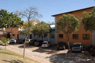 A imagem mostra um edifício de três andares com fachada de cor amarela e laranja, com janelas retangulares escuras. Em primeiro plano, no lado esquerdo, uma árvore de tronco fino, com poucos galhos e folhas verdes, se destaca. O prédio parece ser composto por módulos, com um telhado de duas águas. À direita do prédio, um estacionamento com piso de paralelepípedos e árvores separa o edifício de uma fileira de carros estacionados.