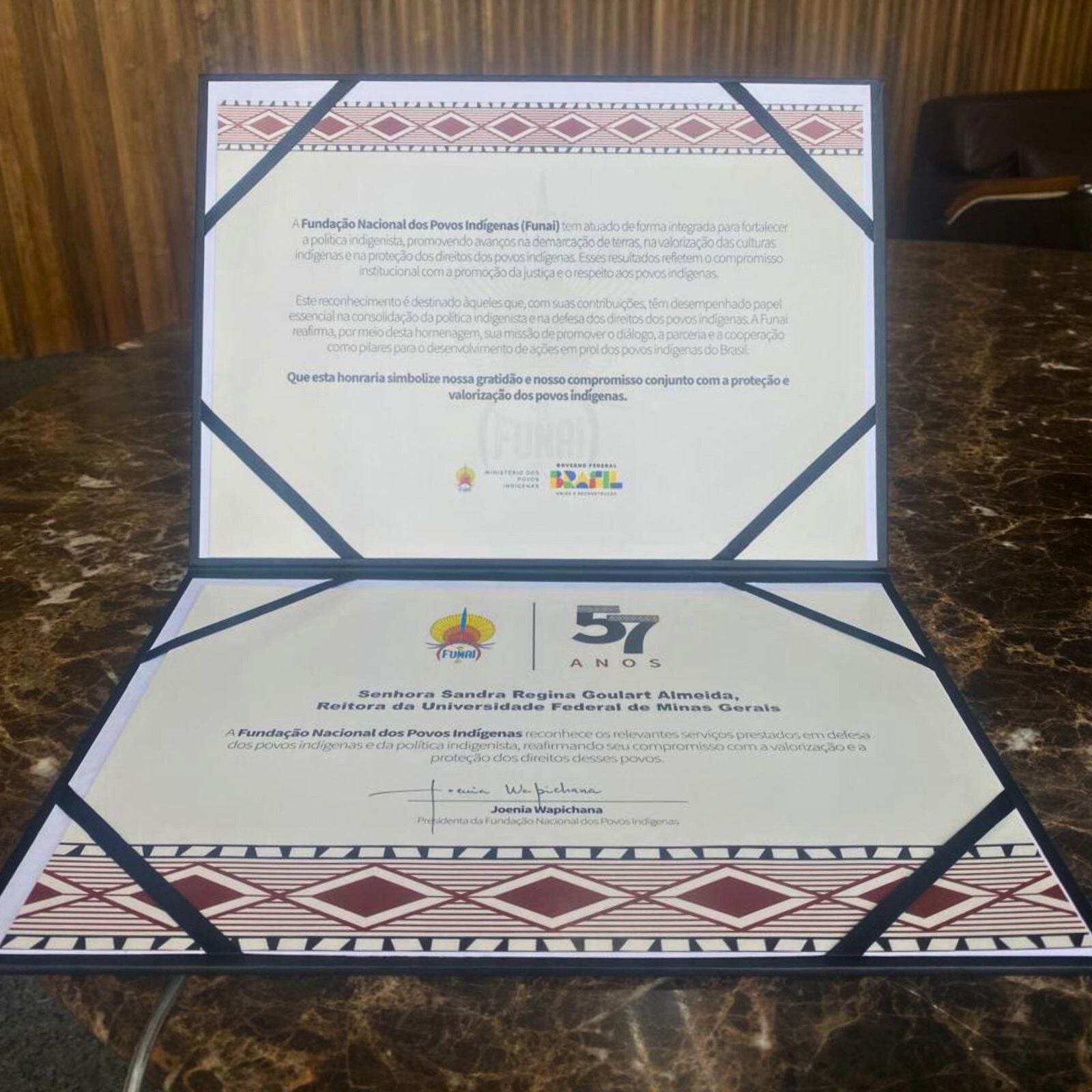 A imagem mostra dois certificados ou diplomas abertos sobre uma superfície escura. Ambos são emoldurados por uma borda decorativa com padrões geométricos em tons de vermelho escuro e branco, e fitas pretas nos cantos superiores. O certificado de cima contém um texto detalhado sobre a atuação da Fundação Nacional dos Povos Indígenas (Funai) e seus objetivos, com o logotipo da Funai e o do governo federal visíveis no meio. O certificado de baixo destaca uma homenagem a "Senhora Sandra Regina Goulart Almeida, Reitora da Universidade Federal de Minas Gerais", com a assinatura de "Joenia Wapichana, Presidenta da Fundação Nacional dos Povos Indígenas". Há também a inscrição "57 ANOS" em destaque.