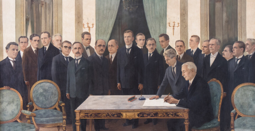 Fotografia histórica em preto e branco, tirada em frente à entrada da antiga Escola de Enfermagem "Carlos Chagas". No topo da imagem, há uma placa grande com o nome da instituição, ladeada por dois símbolos em forma de cruz estilizada. Embaixo da placa, um grupo de aproximadamente quinze pessoas posa para a fotografia, a maioria composta por homens de terno e gravata, muitos deles segurando chapéus nas mãos. Ao centro, destaca-se uma mulher vestida com uniforme branco de enfermeira, incluindo touca e luvas, sorrindo levemente. O grupo está em frente a um prédio de arquitetura clássica, com janelas e sacadas decoradas. Há automóveis antigos parcialmente visíveis nas laterais da imagem.