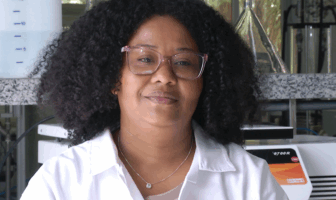 Pesquisadora Rayane Silva, mulher negra com cabelos cacheados na altura dos ombros e usa jaleco branco