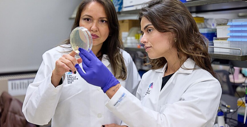 Camila Coelho e Rainna Fantin são duas mulheres vestidas de jaleco branco; ambas seguram um recipiente de vidro ou de plástico em um laboratório