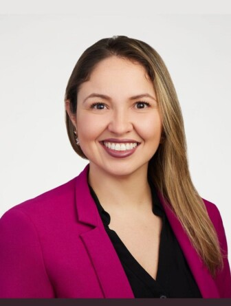 Camila Coelho é uma mulher de cabelos castanhos e usa uma roupa vermelha. Ela sorri na foto.