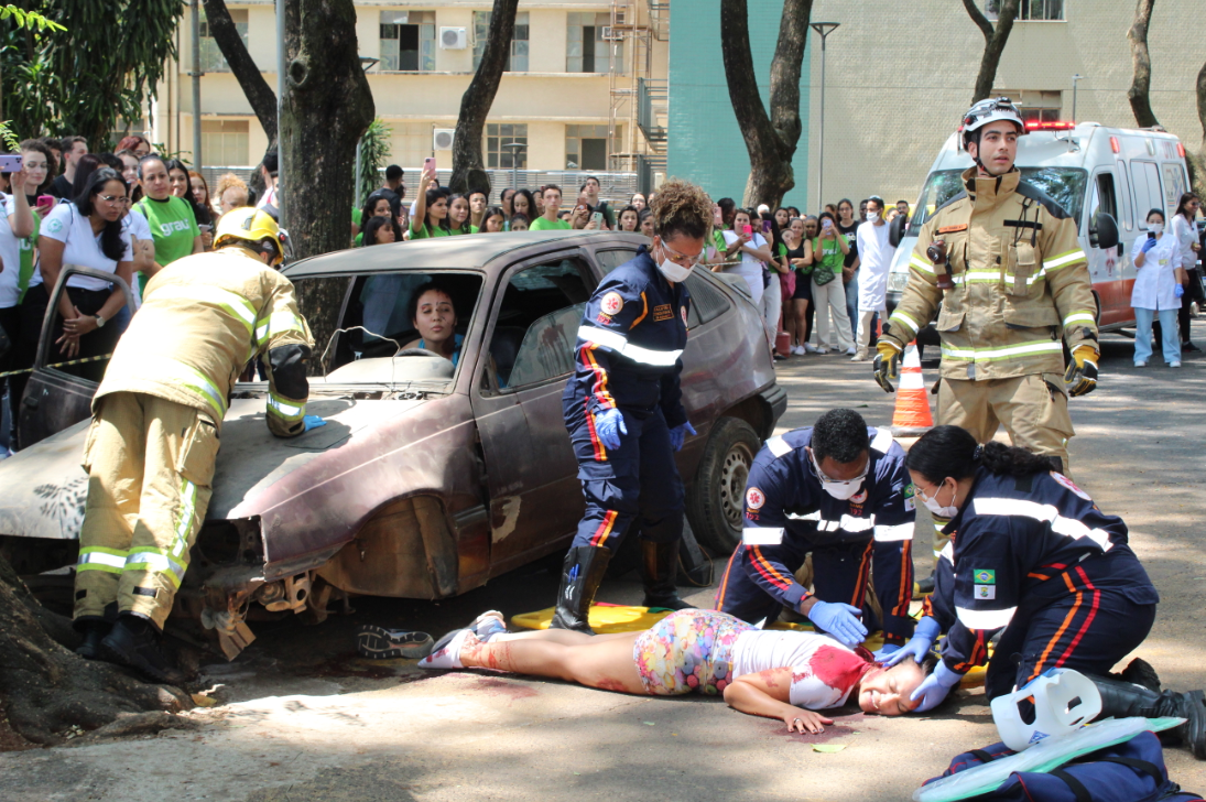 A imagem mostra uma simulação de acidente com várias pessoas envolvidas. Em primeiro plano, uma figura feminina está deitada no chão, com a cabeça apoiada no ombro de um socorrista. Ela veste uma blusa branca e shorts estampados. Três socorristas, usando uniformes azuis escuros com detalhes refletivos, luvas e capacetes, estão prestando atendimento à vítima no chão. Um carro amassado e parcialmente destruído está no centro da cena. No banco do motorista do carro, uma pessoa com semblante preocupado pode ser vista. Outros dois socorristas, vestidos com uniformes amarelos e capacetes, estão próximos ao veículo, um deles olhando para a pessoa no carro. Ao fundo, uma ambulância branca e um grupo de observadores formam uma multidão. O cenário inclui árvores e edifícios ao fundo.