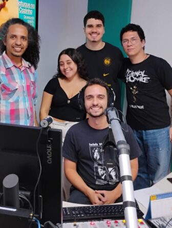 Bruno Vasconcelos, de camisa xadrex colorida, ao lado de Isabela Fernandes, Luiz de Andrade, Hugo Rezende e Hugo Oliveira, todos de blusa preta, no estúdio de rádio, com microfone e computador