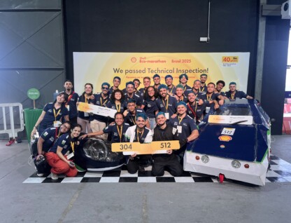 Na frente do banner amarelo da competição estão 28 alunos, todos com uniforme azul marinho e medalhas penduradas no pescoço. Eles estão ao lado de dois prótotipos de carros