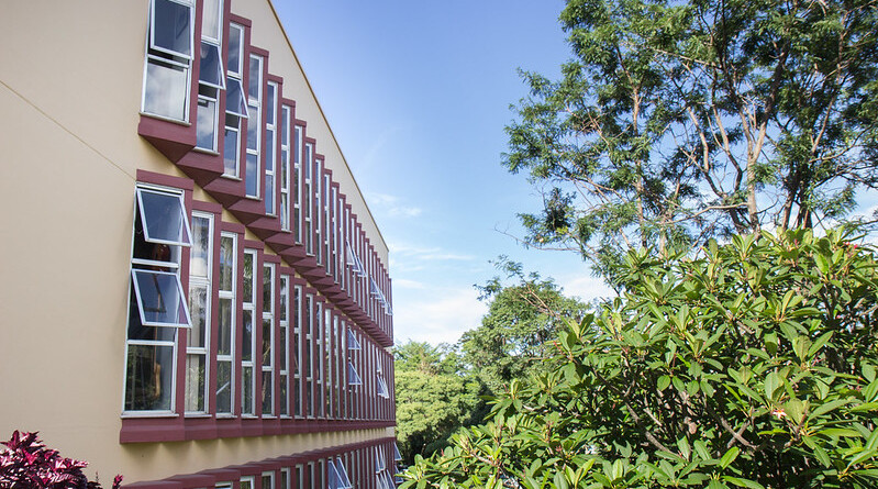 Fachada lateral da Escola de Música, cor laranja claro, com várias janelas com tom de vermelho escuro; ao lado, há árvores