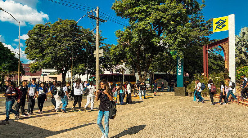 Estudantes andando em frente à Praça de Serviços; à direita, placas com logo de bancos; ao fundo, árvores e dois ônibus