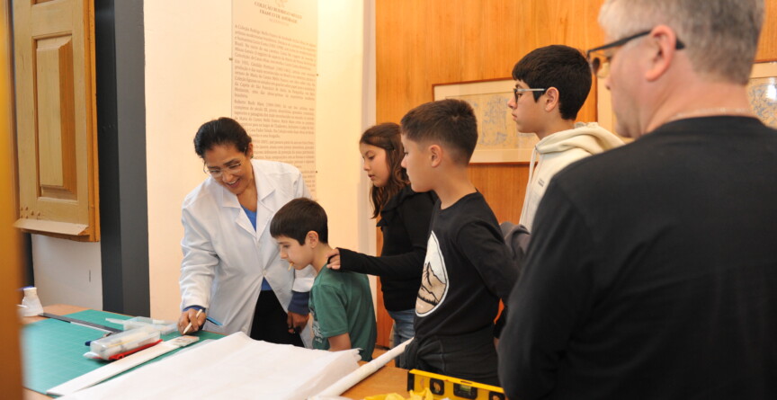 Crianças visitam exposição no Museu Padre Toledo; uma mulher de  jaleco branco explica detalhes da mostra. À direita, um homem grisalho, de óculos e blusa preta observa a movimentação