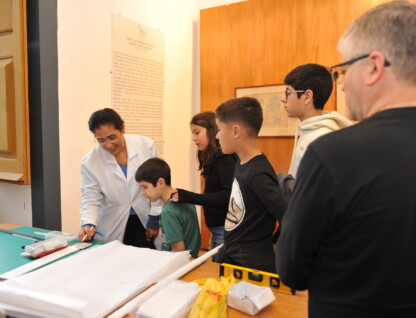 Grupo de crianças visita a exposição acompanhado de uma mulher de roupa branca; atrás, um homem de roupa escura, grisalho e de óculos observa a movimentação